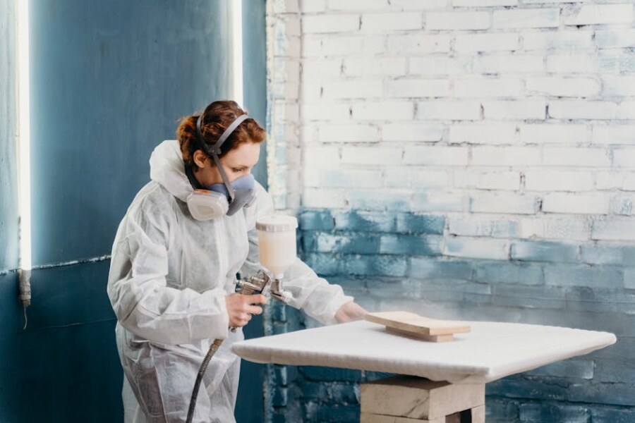 A female artisan wearing protective gear spray paints a wooden object indoors.